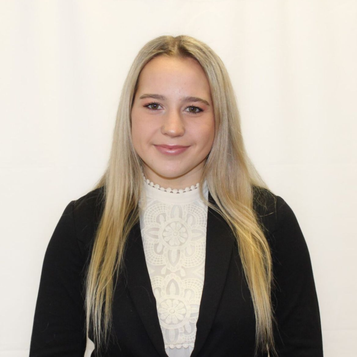 Woman with long blonde hair, wearing a black blazer over a white lace top, smiling. White background.