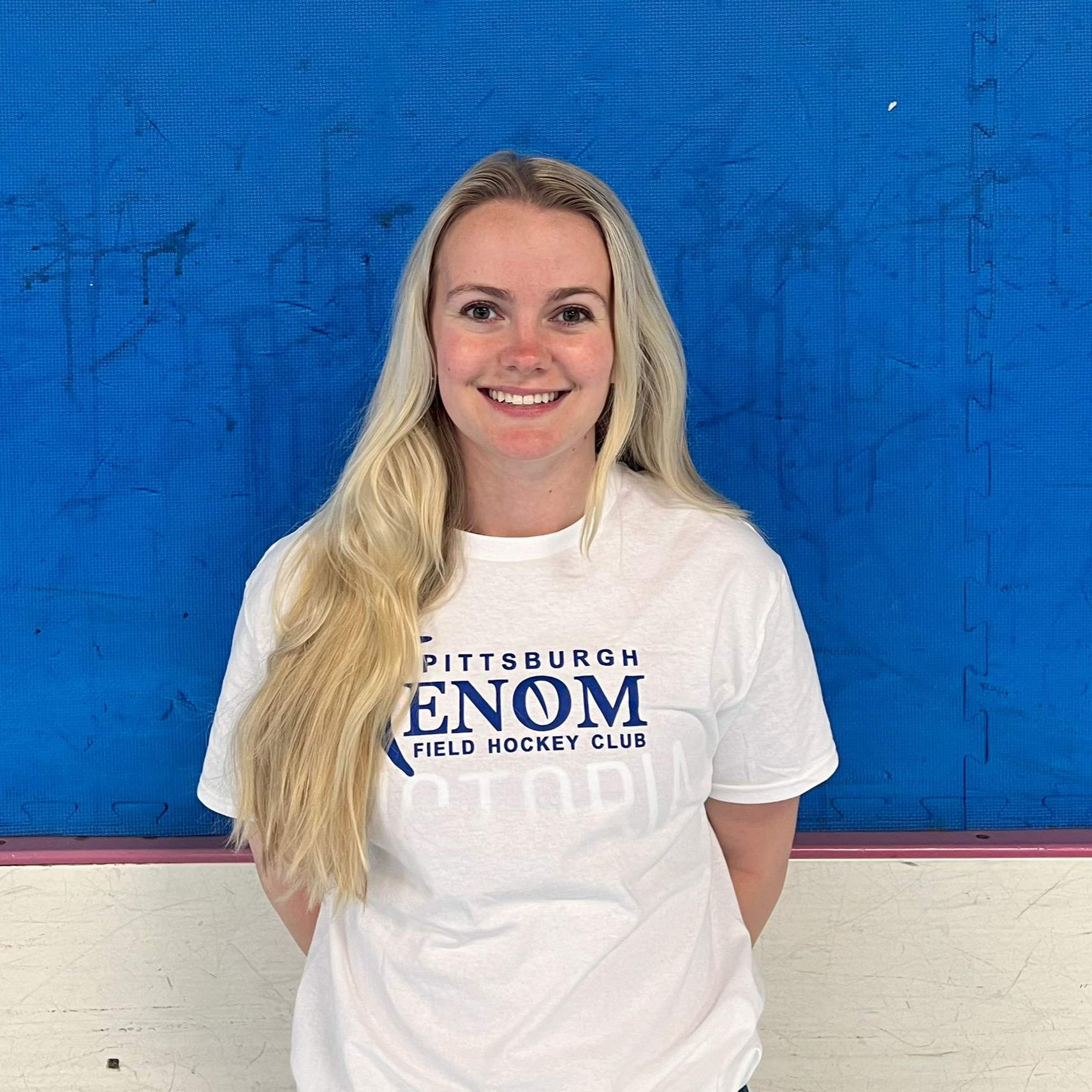 Blonde woman in white t-shirt, smiling, stands in front of a blue background with a Pittsburgh Venom Field Hockey Club logo.
