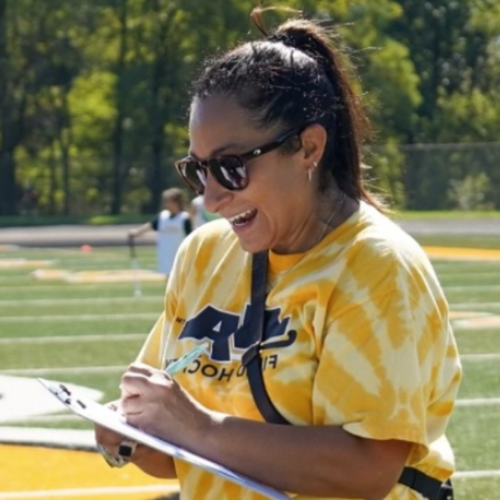 Woman in sunglasses, yellow tie-dye shirt, ponytail, smiles while writing on a clipboard outdoors on a sports field.