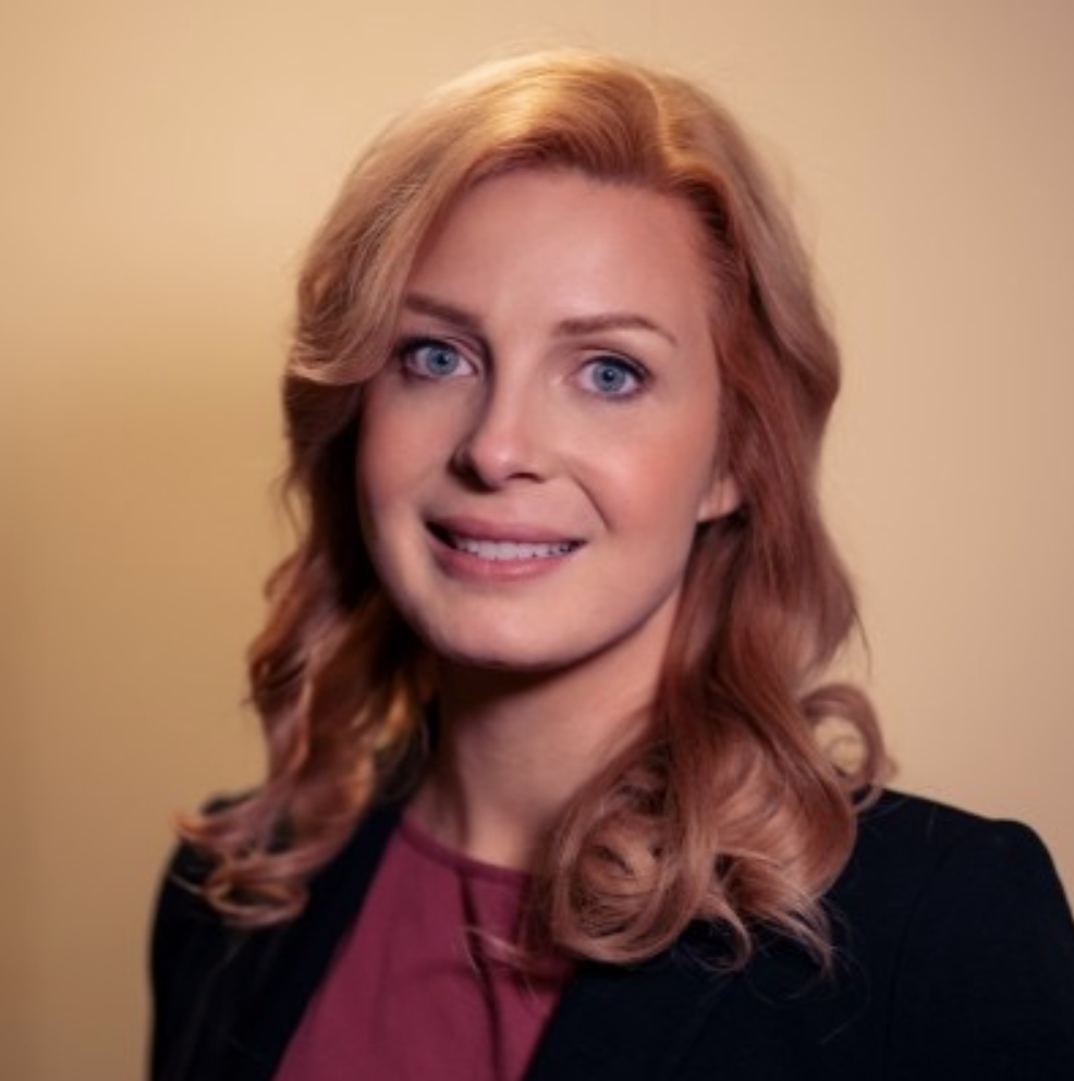 Woman with reddish-blonde wavy hair smiles, wearing a black blazer and maroon top, against a yellow background.