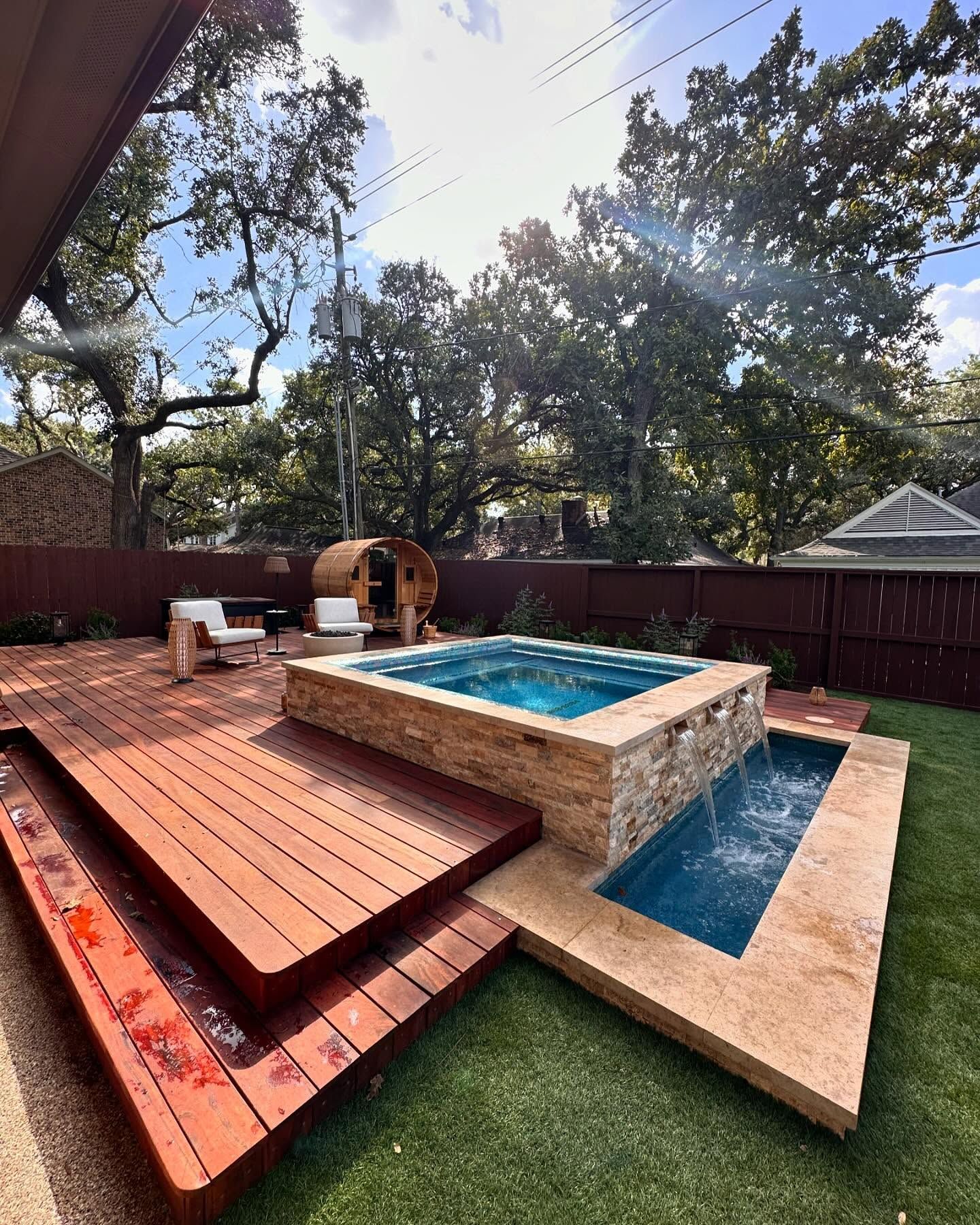 A backyard with a wooden deck, a hot tub with a waterfall, and a sauna, set against a backdrop of trees and blue sky.