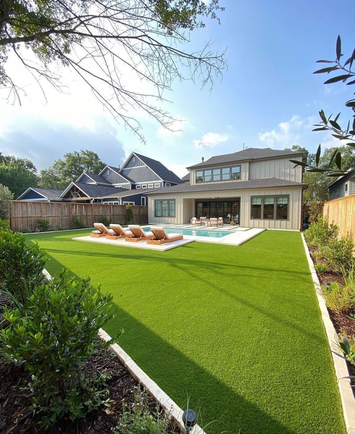 Backyard with a pool, lawn, and two-story house.  Green grass, lounge chairs by the pool, and a wooden fence.