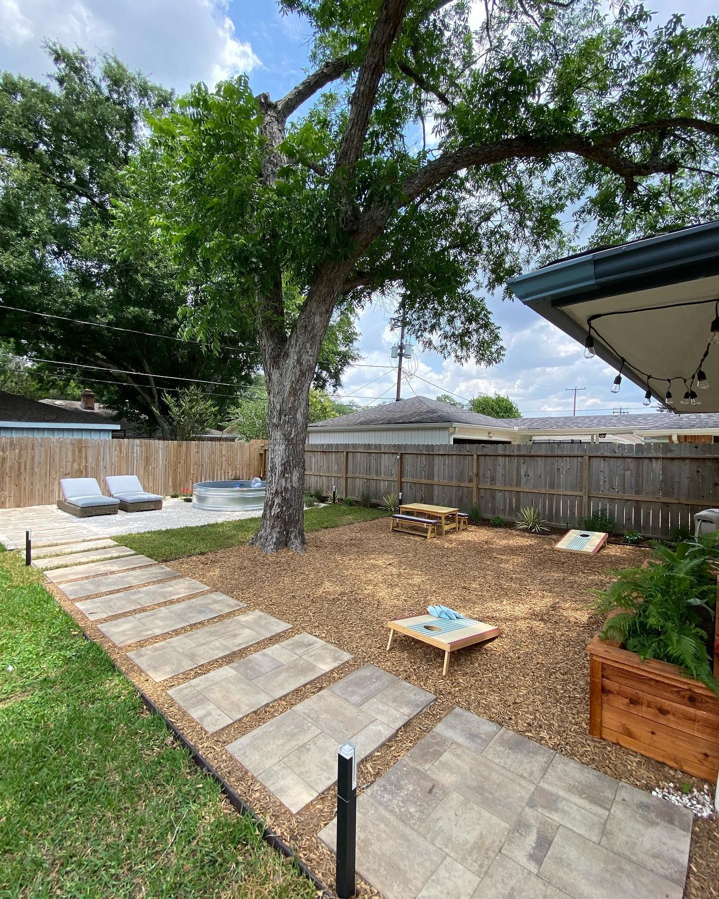 Backyard with stone path, lounge chairs, hot tub, picnic table, cornhole, and large tree.