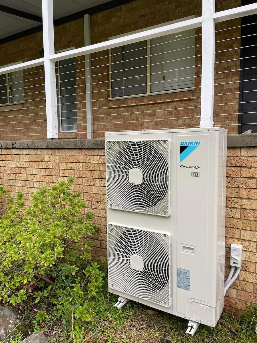 Outdoor Wall-mounted Air Conditioner Unit Beside a Brick Building Window and Bushes — Deltech HVACR in Georgetown, NSW