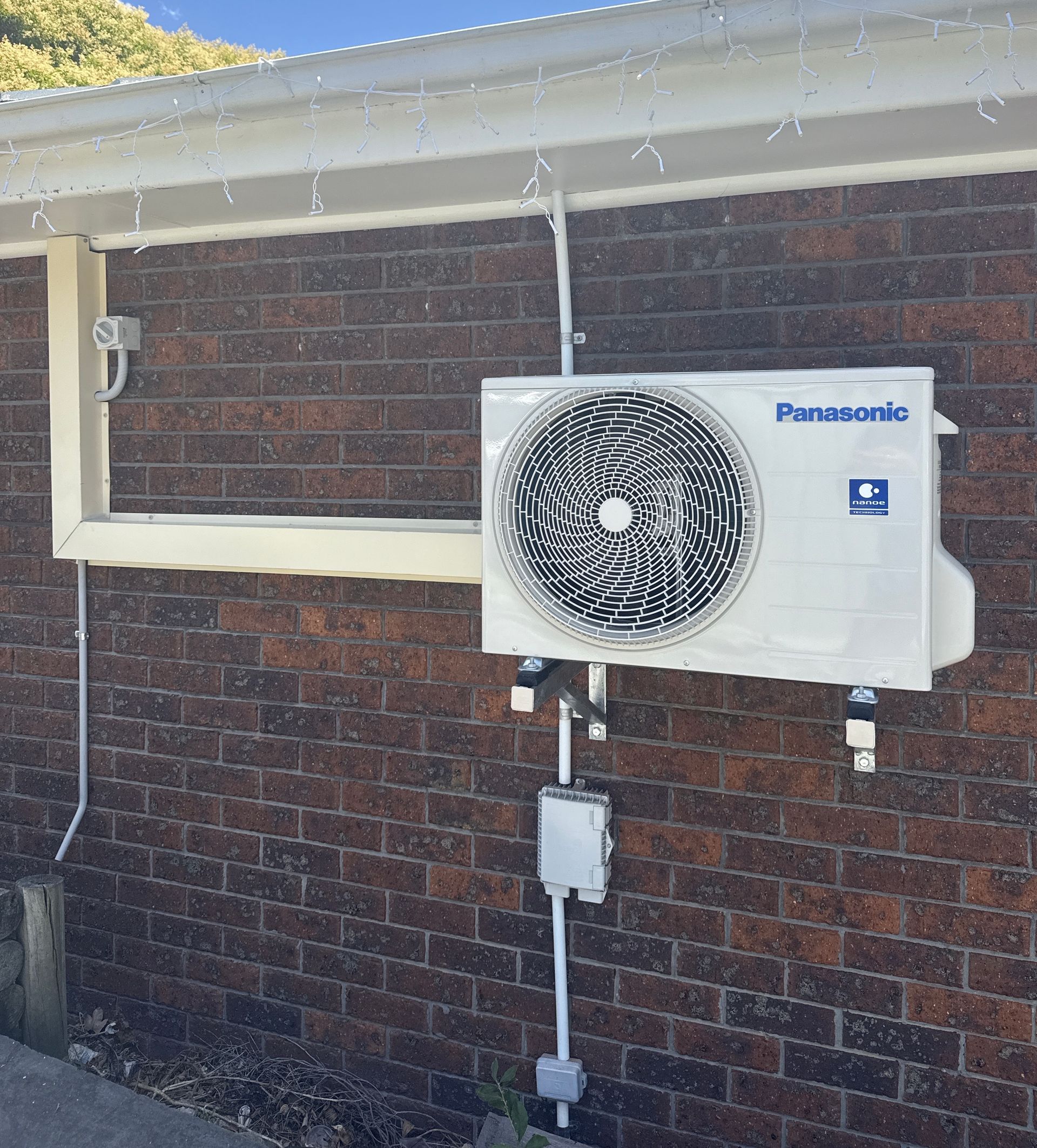 Outdoor Panasonic AC unit mounted on a brick wall beside a white utility conduit, with water dripping overhead — Deltech HVACR in Georgetown, NSW