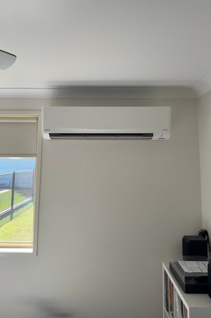 White wall with a mounted air conditioner above a window and a desk in the corner — Deltech HVACR in Georgetown, NSW