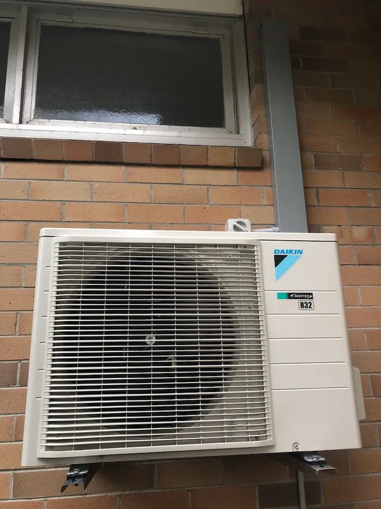 Wall-mounted Outdoor Air Conditioner Unit Beneath a Window on a Brick Wall — Deltech HVACR in Georgetown, NSW
