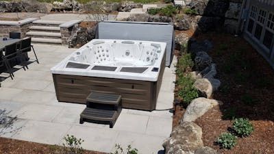 Hot tub on a stone patio next to a garden bed and steps, covered by a gray cover.
