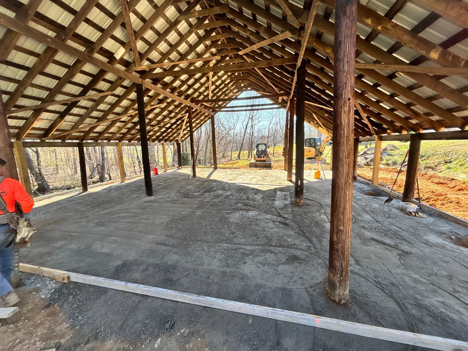 Open-air barn under construction with gravel floor, wooden beams, and metal roof. Construction worker on the left.