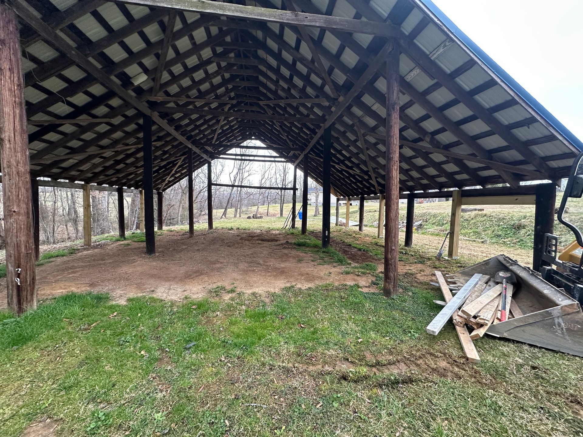 Open-sided wooden structure with a metal roof. Inside, the ground is covered in tan material. Surrounded by green grass.