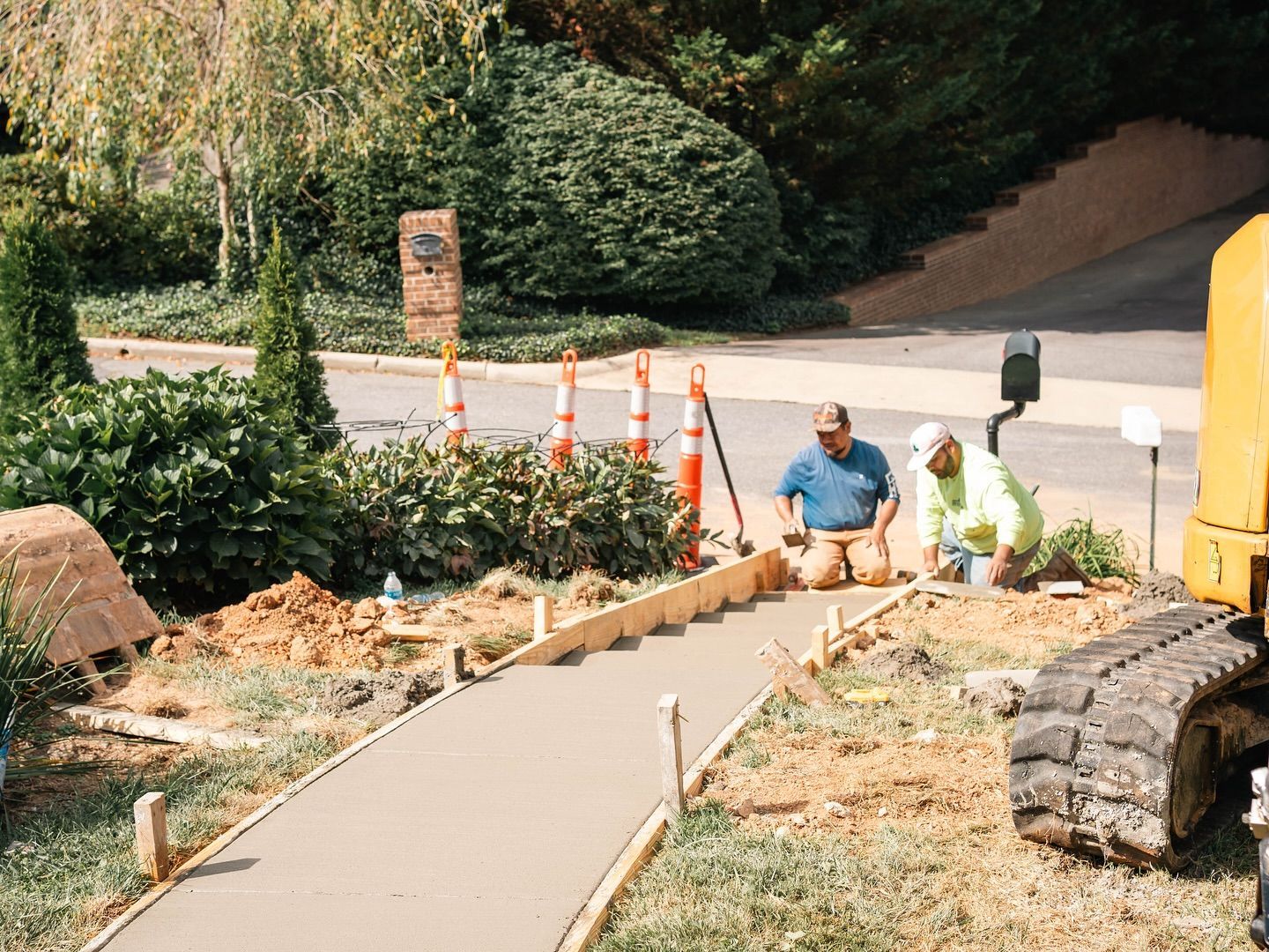 Two workers pouring concrete for a sidewalk, framed by wooden boards, with a small excavator nearby.