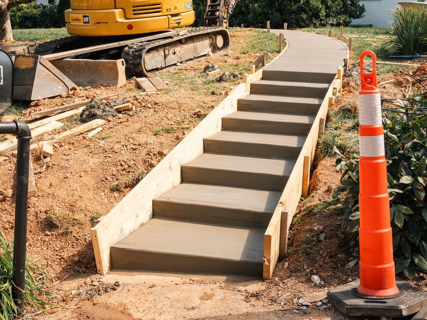 Newly poured concrete steps in wooden forms, next to orange traffic cone and excavator.