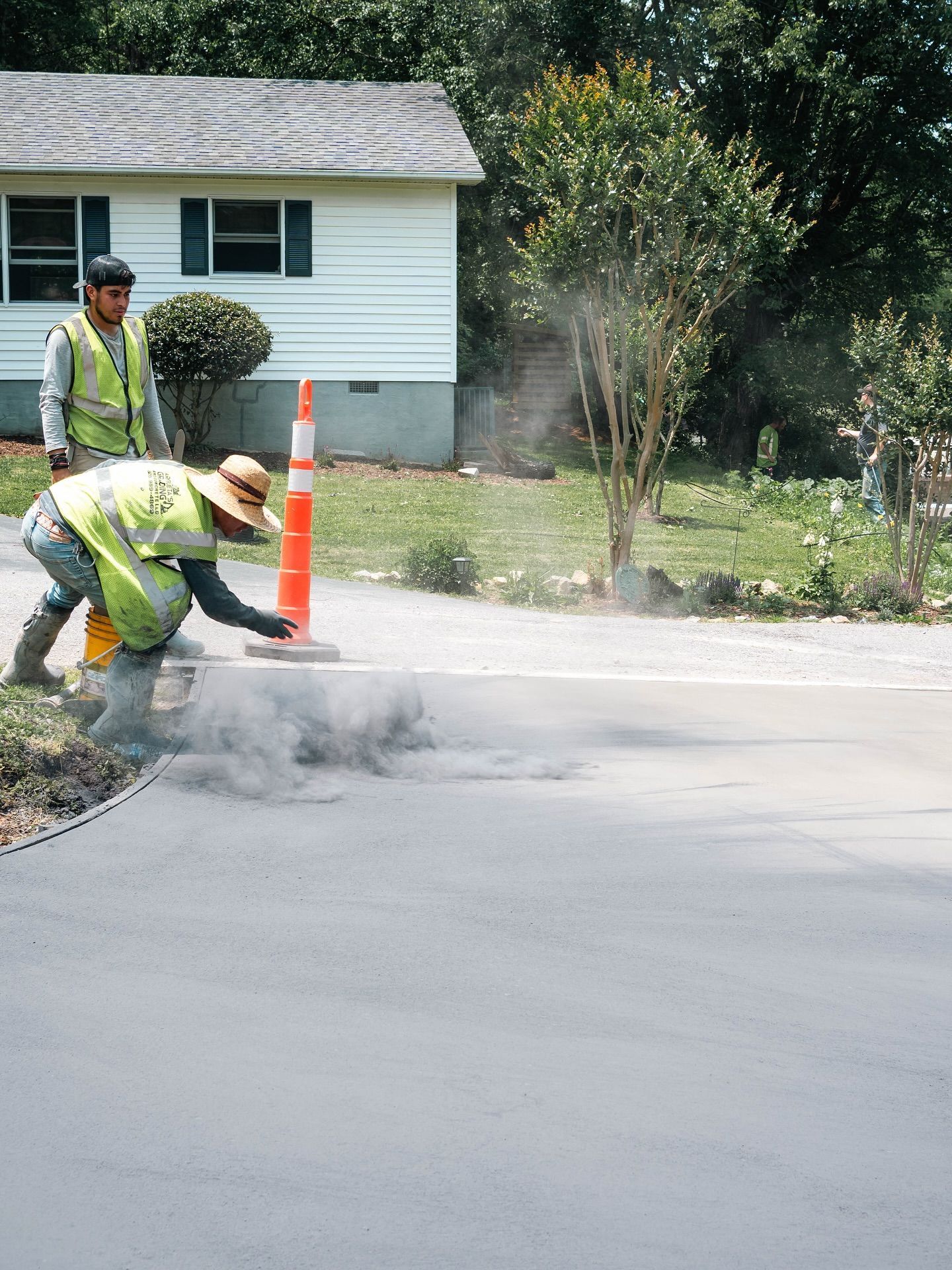 Two workers in safety vests resurfacing a road. One kneels, applying material, while the other observes near a traffic cone.
