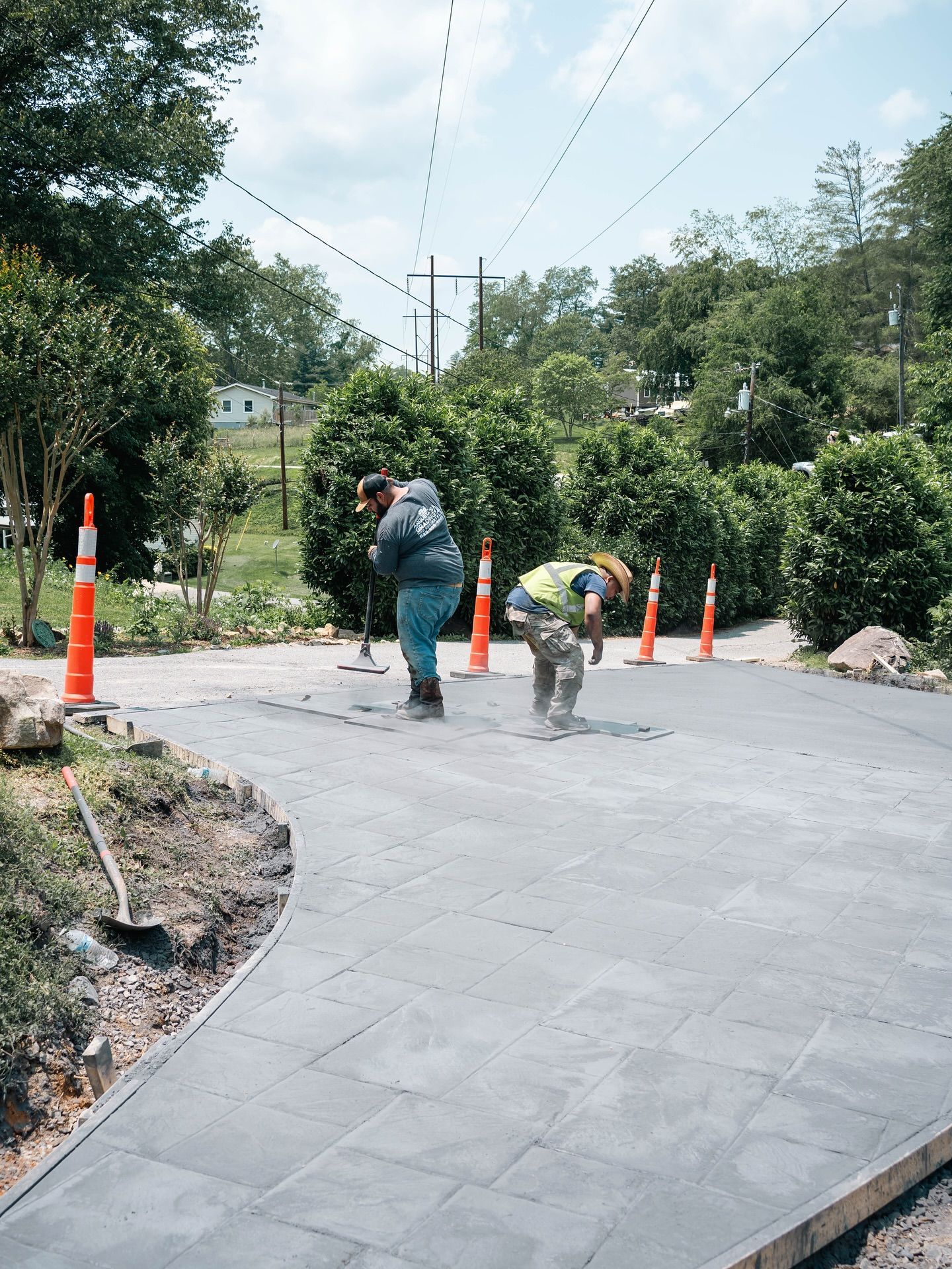 Two workers installing pavers outdoors, near traffic cones and shrubbery, on a sunny day.