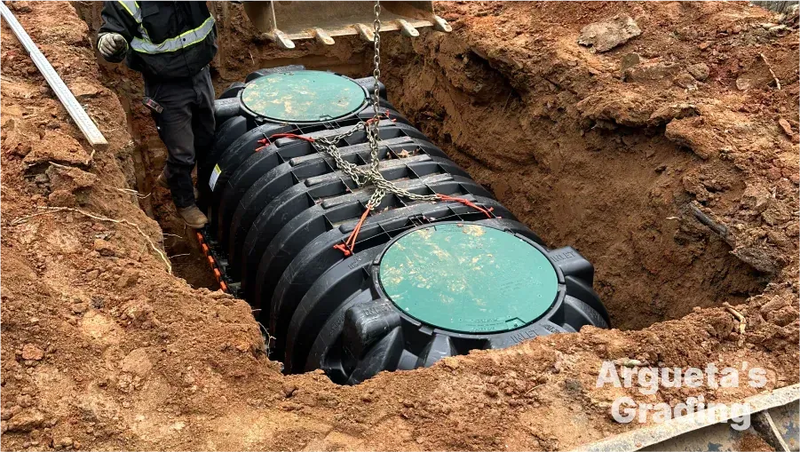 Septic tank being lowered into an excavated trench by a construction worker using chains and machinery.