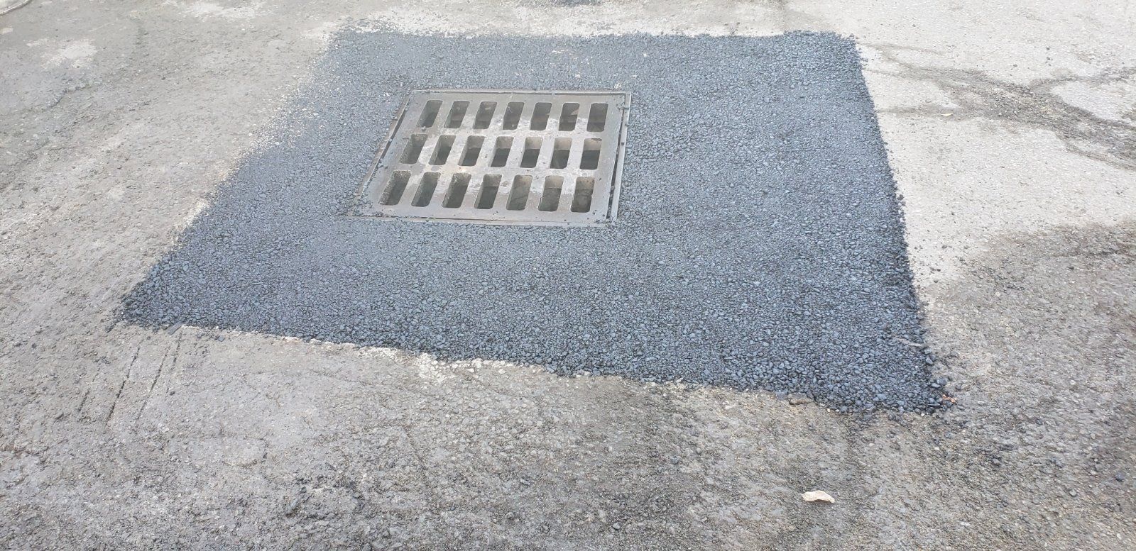 Square drainage grate surrounded by gravel on a concrete surface.