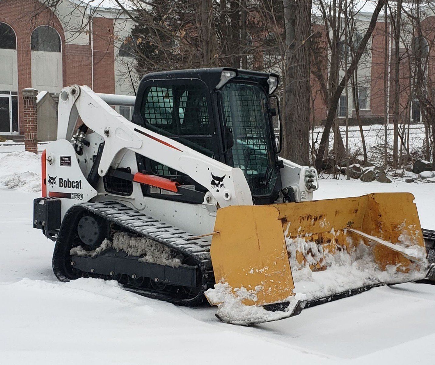 A white Bobcat compact track loader with a yellow snowplow blade clears snow in front of a building.