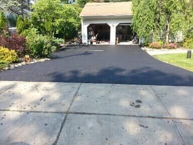 A driveway with a freshly paved black asphalt section leading to a garage. The driveway borders grass and shrubs.