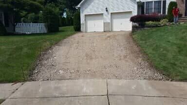 Gravel driveway in front of a two-car garage. The driveway is rough and uneven, transitioning to a paved sidewalk.