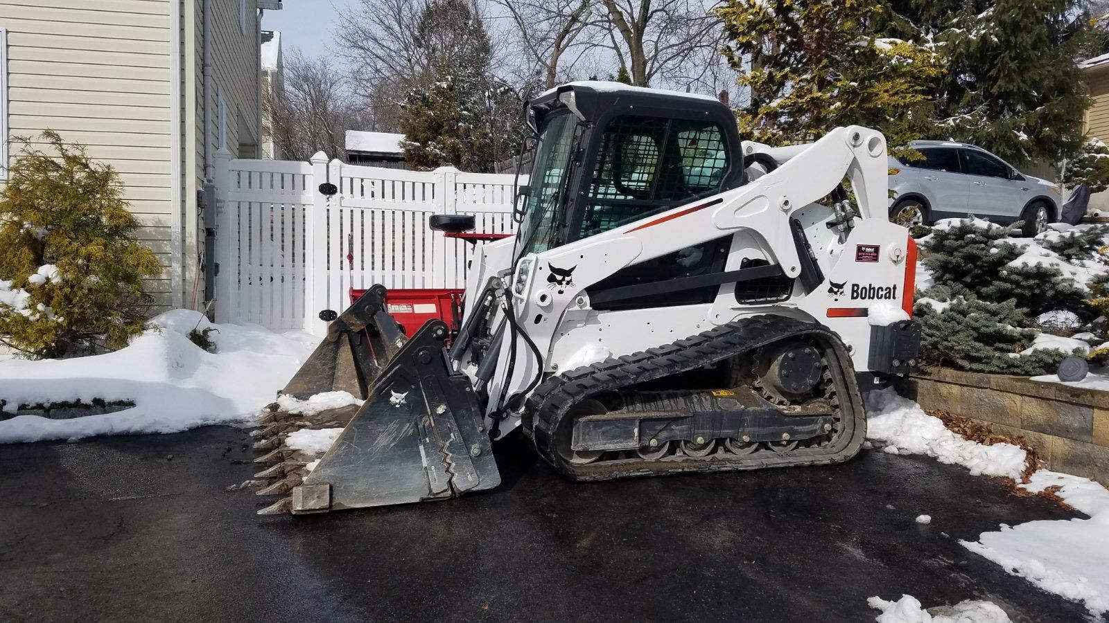 White Bobcat track loader clearing snow from a dark driveway near a white fence and a partially visible car.