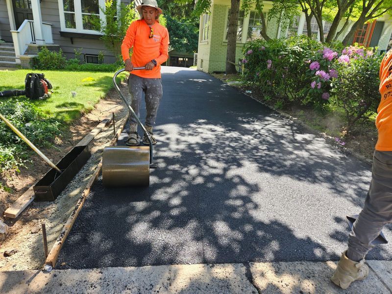 Workers paving a driveway with asphalt; one uses a roller, others stand nearby.