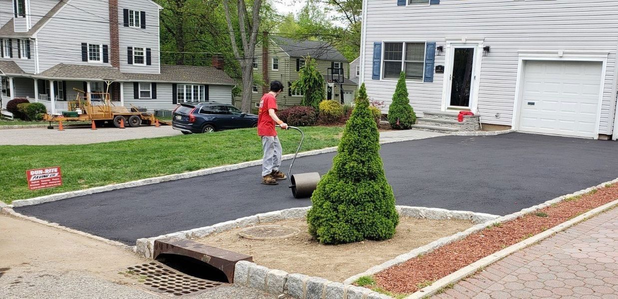 A person rolls a large cylinder on a newly paved driveway near a house with green landscaping.
