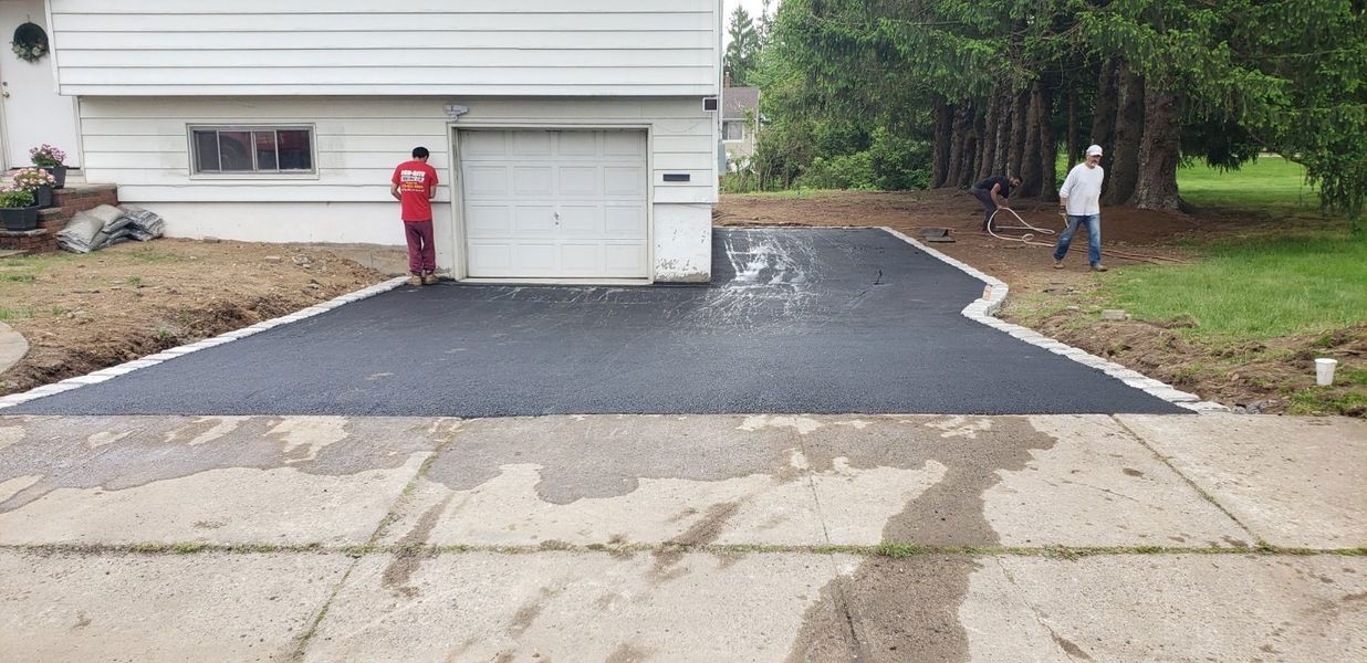 Freshly paved driveway next to a white house. Two workers are present; one by the garage door, another near a tree.