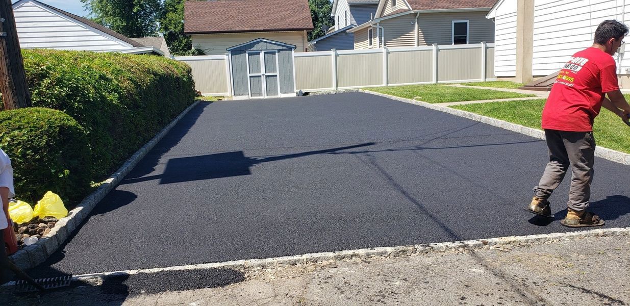 A person in a red shirt paving a driveway with fresh black asphalt, in a residential neighborhood.