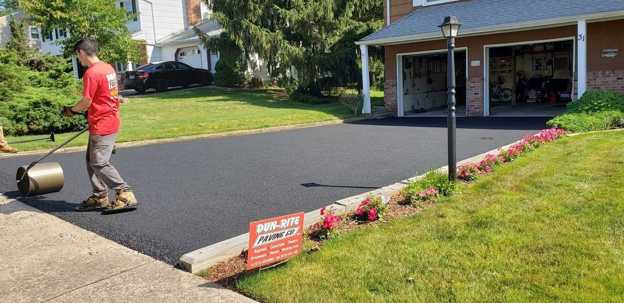 A worker uses a roller on a newly paved driveway. A house with a garage is in the background.