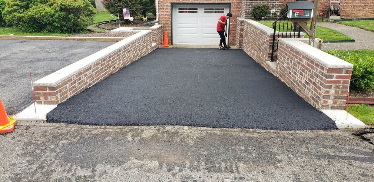 A driveway is being paved with fresh asphalt. Brick walls border the asphalt as a worker applies it.