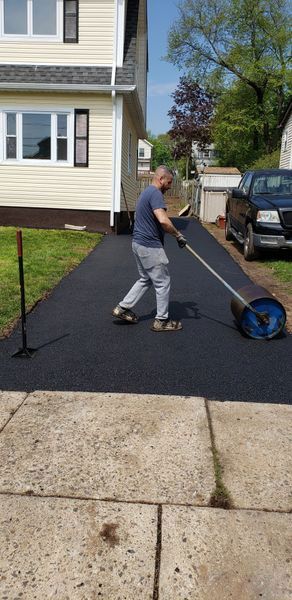 A man rolling asphalt on a driveway near a two-story house. The driveway is black; the house is beige.