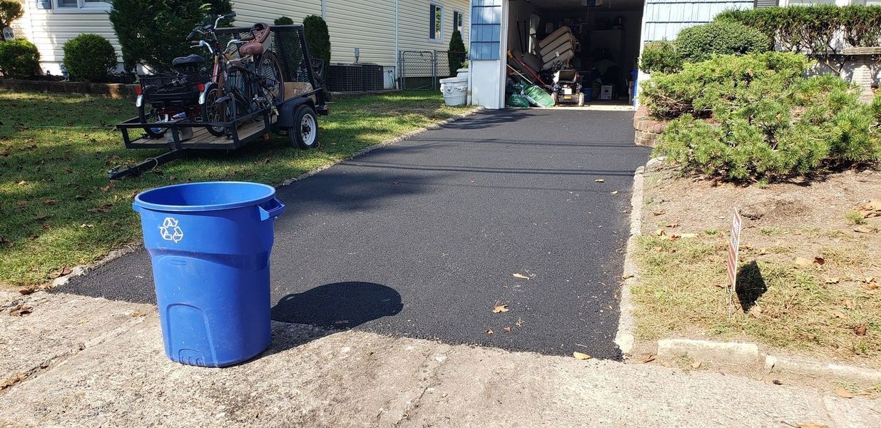 A blue recycling bin sits in front of a newly paved asphalt driveway, near a garage and some landscaping.