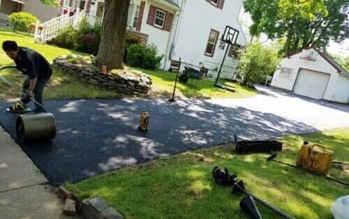 A person rolling out fresh asphalt on a driveway in front of a house on a sunny day.