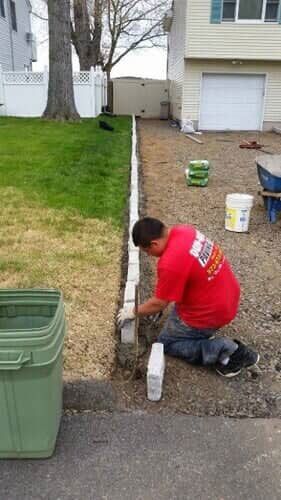 A person in a red shirt kneels, installing edging stones along a driveway. They work next to a green trash bin and a lawn.