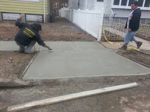 Two men paving a concrete sidewalk. One kneels, smoothing, the other walks nearby.