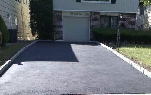 Asphalt driveway leading to a garage with a brick facade; green lawn and bushes on either side.