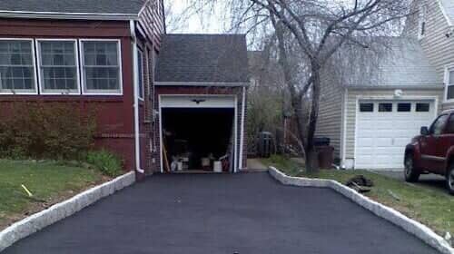 Asphalt driveway leading to open garage between two houses; a red car is parked on the right.