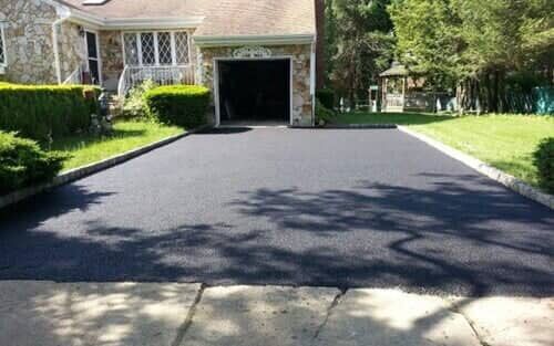 A newly paved asphalt driveway in front of a house with a garage, framed by grass and shrubs.
