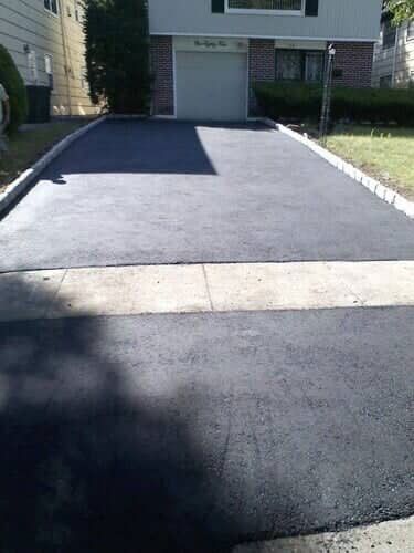 Newly paved black asphalt driveway leading to a gray garage with a house in the background.