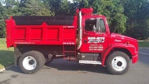 Red dump truck parked on pavement, advertising a business. The truck has white wheels and a side ladder.
