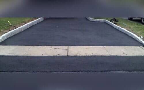 Asphalt driveway with a concrete border, leading from a sidewalk to a green lawn. The driveway has a dark, smooth texture.