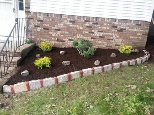 Triangular garden bed with a brick border, filled with dark mulch and various plants in front of a brick wall.