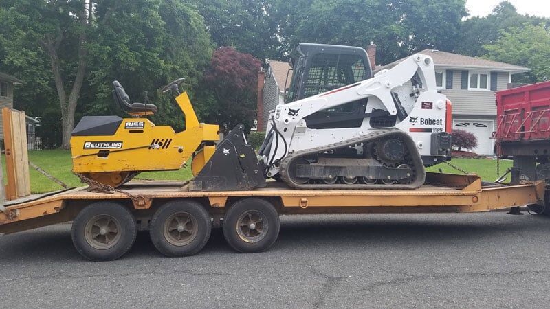 A yellow roller and white Bobcat track loader on a trailer, parked on a residential street.