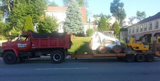 Red dump truck towing a flatbed trailer carrying a white skid steer and yellow