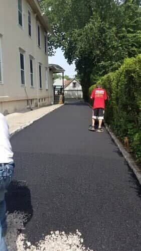 Workers paving a long, narrow asphalt driveway next to a building. One worker rolls the asphalt.