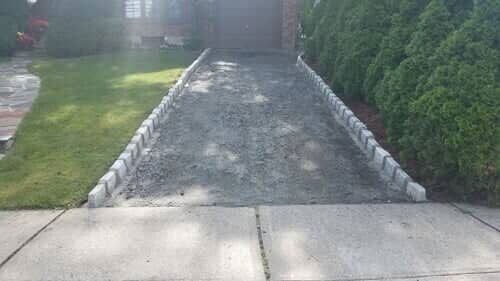 Driveway with gravel, flanked by concrete pavers and a strip of grass on one side, a hedge on the other, leading to a garage.