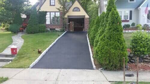 A paved driveway leads to a house with an open garage. Tall, green trees line the right side.