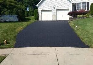 A freshly paved black asphalt driveway leading to a white house with a two-car garage, surrounded by green grass.