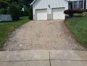 A gravel driveway leading to a white house with a two-car garage; green lawn surrounds.