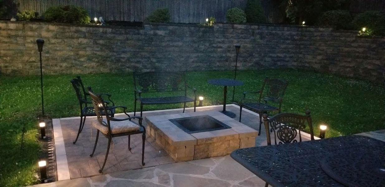 A nighttime backyard patio with a fire pit and seating, illuminated by path lights.